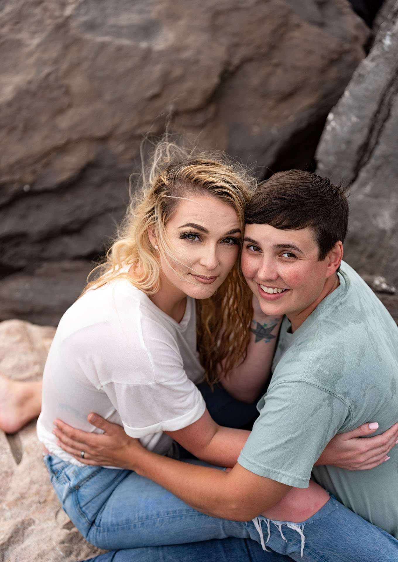 Two women embracing and smiling on rocks at St. Andrews State Park, one with windswept blonde hair, one with short dark hair