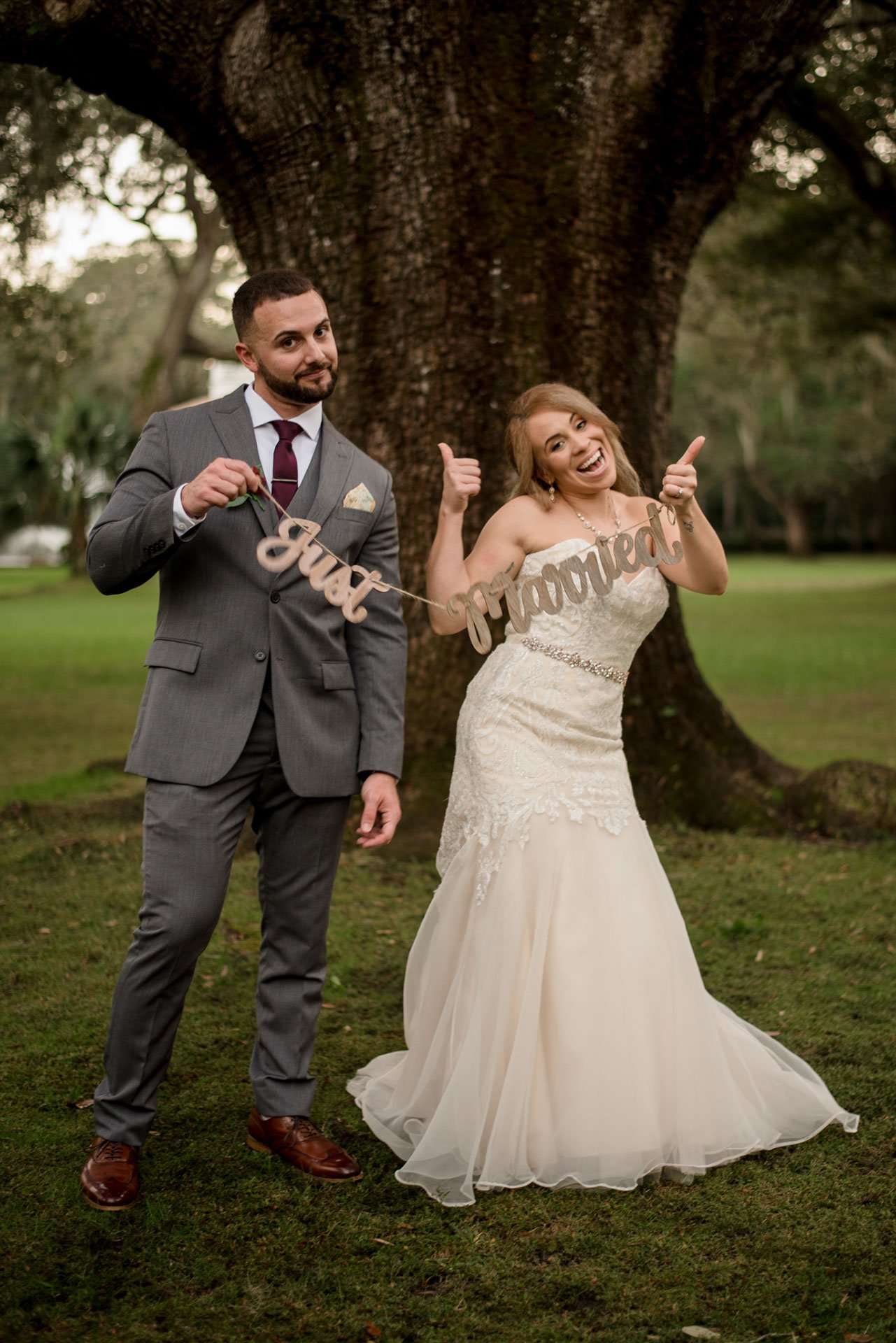 Bride and groom holding a Just Married sign, bride giving thumbs up, in front of a large oak tree at Eden Gardens State Park