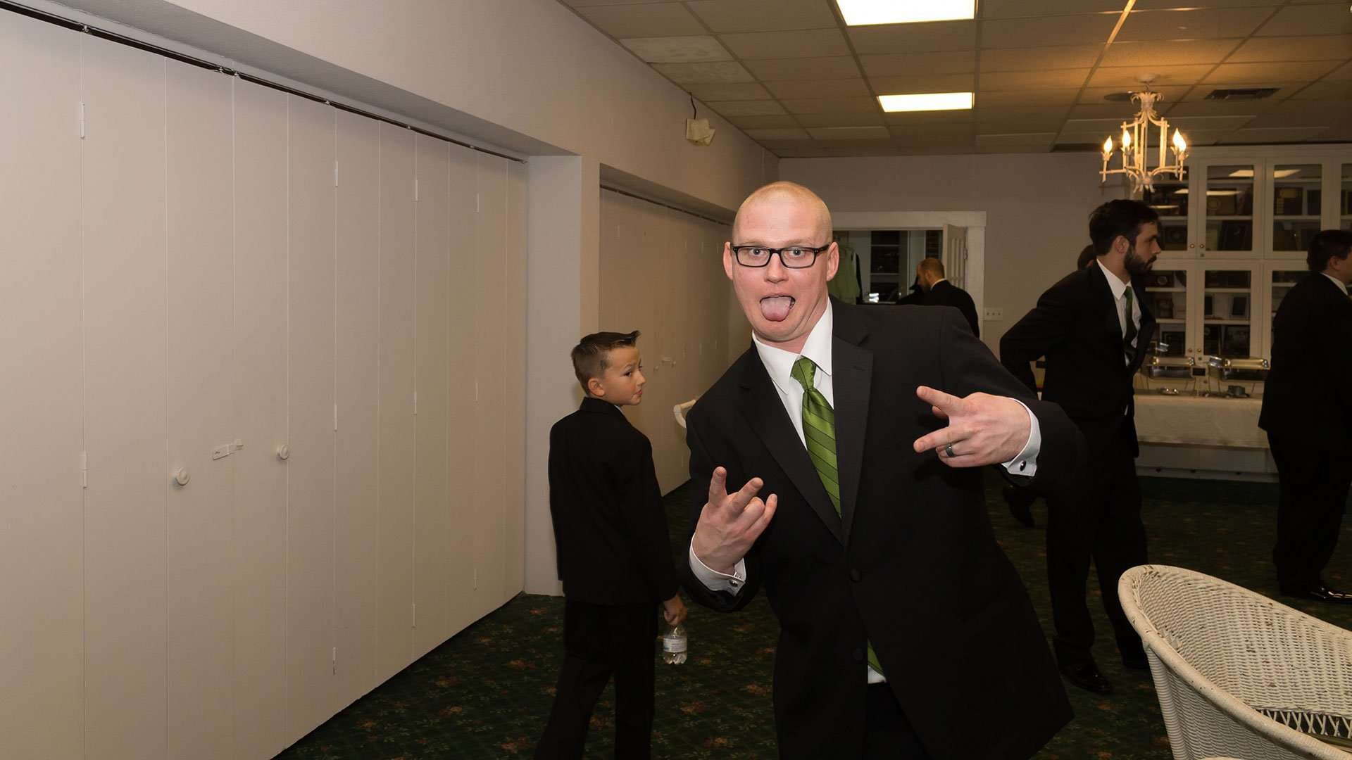 Bald man in glasses, black suit and green tie sticks out his tongue and flashes hand signs at a reception