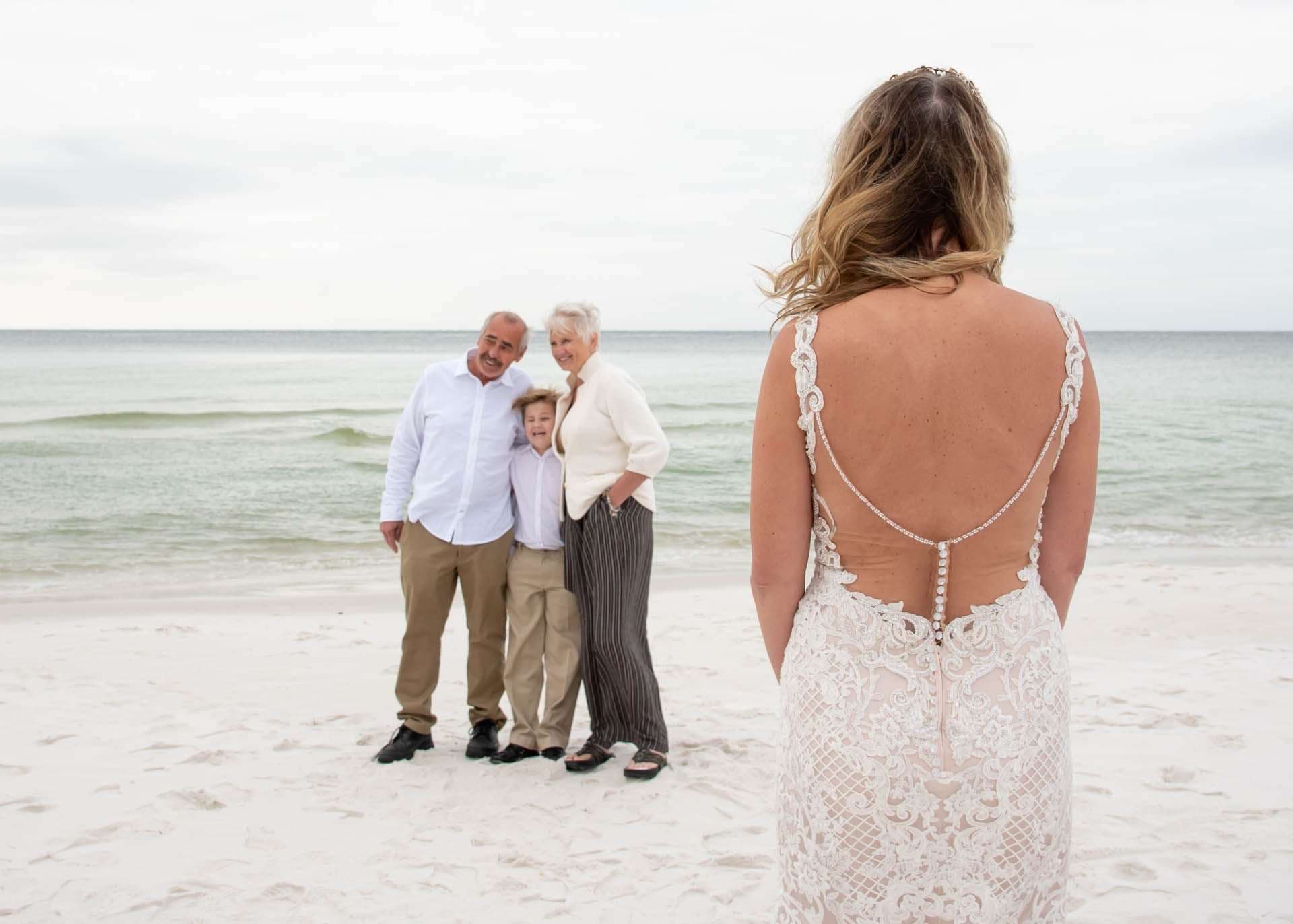 Bride in a lace low-back gown seen from behind, facing an older man, older woman and boy posing on the beach